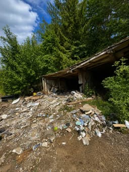 Der Vorher-Zustand einer Räumung. Vor einem alten, baufälligen Holzschuppen am Waldrand liegt ein massiver Berg aus Bauschutt, Sperrmüll und Unrat. Zu sehen sind unter anderem herausgerissene Holzlatten, Rigips- oder Putzplatten, Plastikfolien, alte Tapetenreste und loser Müll, der den Weg komplett blockiert.