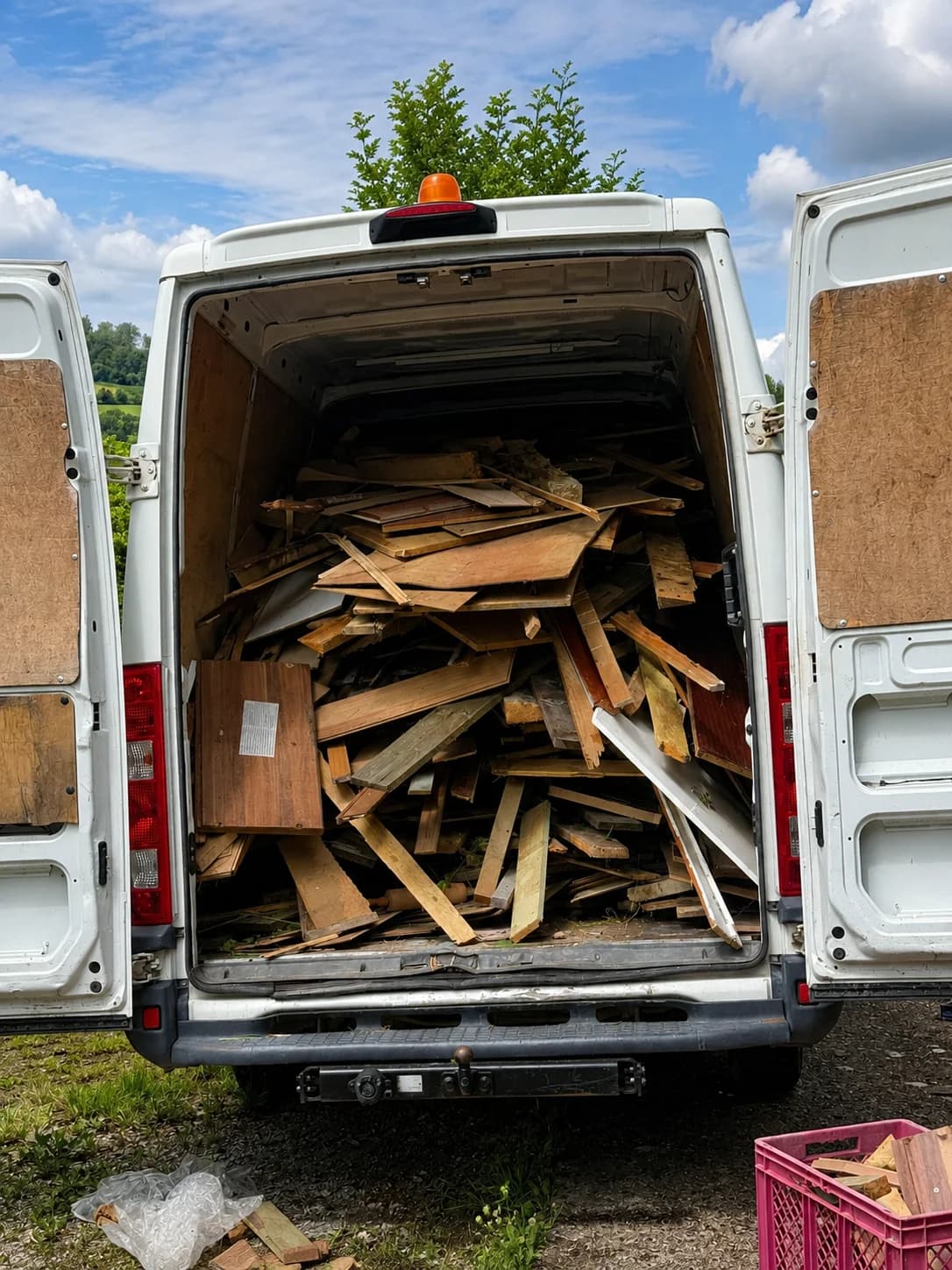 Die Rückansicht des weißen Transporters mit geöffneten Hecktüren. Der Laderaum ist fachgerecht und bis oben hin vollgestapelt mit losem Altholz für die Entsorgung. Zu sehen sind alte Bretter, Spanplatten, Holzleisten und Furnierholz. Auf dem Boden vor dem Auto steht eine rote Klappkiste.